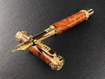 An Artisan Hand Turned Exhibition Grade Exotic Red Mallee Fountain Pen On Red Gold Hardware. Each Finial Has a Rampant Lion Hold A Fountain Pen Engraved. The red gold center band has a series of specifically designed H’s, to represent Highlander. This same band is at the very top of the pen and at thethebottom of the pen where the hardware begins. The clip has a series of these H’s horizontally down it.
The nib is in Red Gold as well, and it is a JOWO #6 Medium.