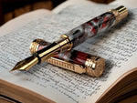 A Red Gold Fountain Pen With Red Gold JOWO #6 Nib. The Body is hand crafted with red, black, and white pearl resins.
The Custom Hardware is Intricately Engraved With A Rampant Lion, which adorns both the cap and finial. The Center Band Showcases A Series Of Understated Yet Distinguished Artist Designed “H” . The Fountain pen is sitting on an old manuscript with the body of the pen resting on the cap.